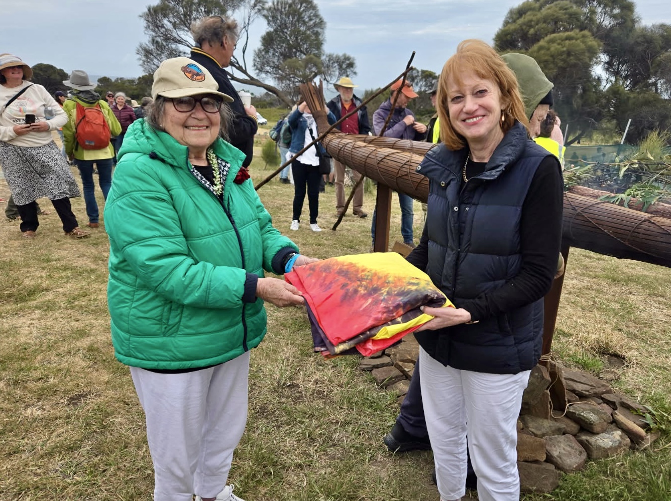 Two women stand together outdoors in front of a Tasmanian Aboriginal Canoe. They are holding a piece of colourful cloth between them and smiling at the camera.