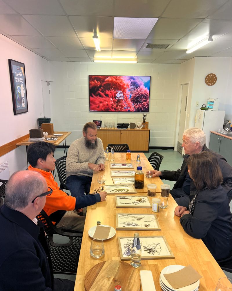 The Governor and Dr Walters sit indoors at a table with three men, looking at seaweed samples