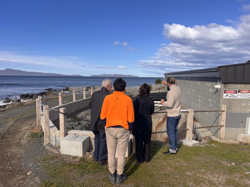 The Governor and Dr Walters stand outdoors with two men next to a building, looking out at the sea in the distance