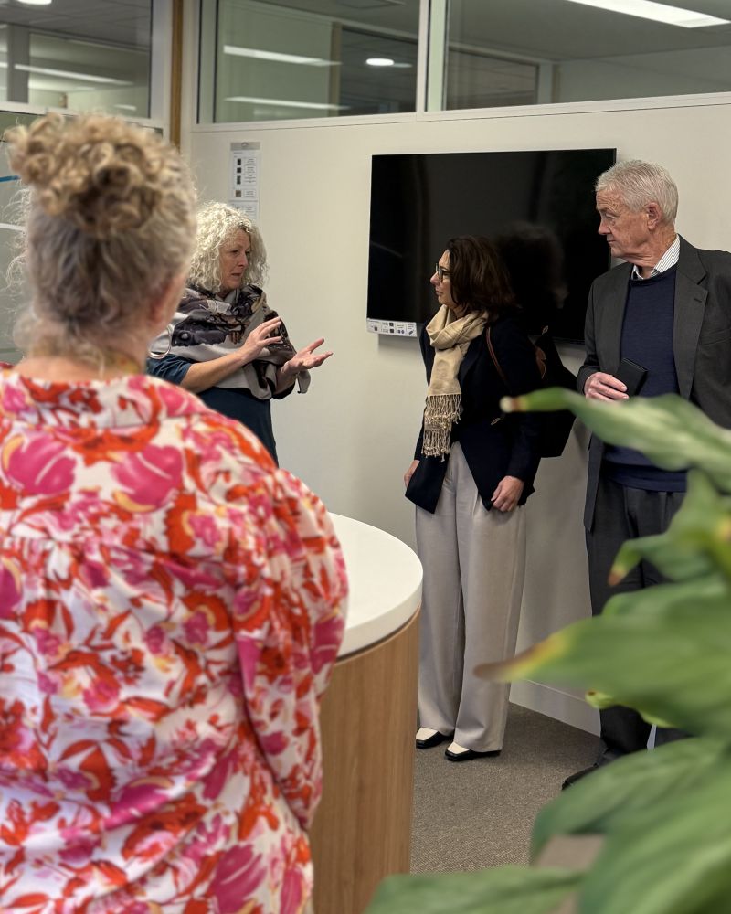 The Governor and Dr Walters stand indoors in a reception area listening to a woman speak