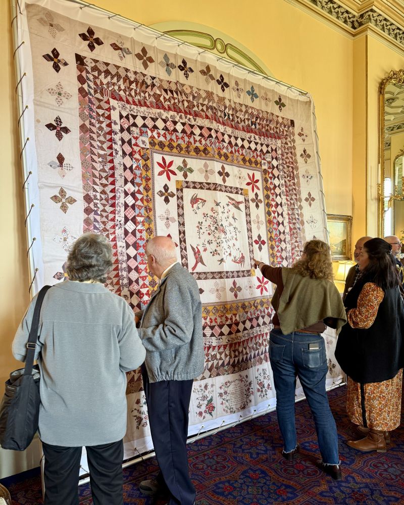 Five people stand in the Government House drawing room. They have their backs to the camera as they admire a large colourful quilt on display