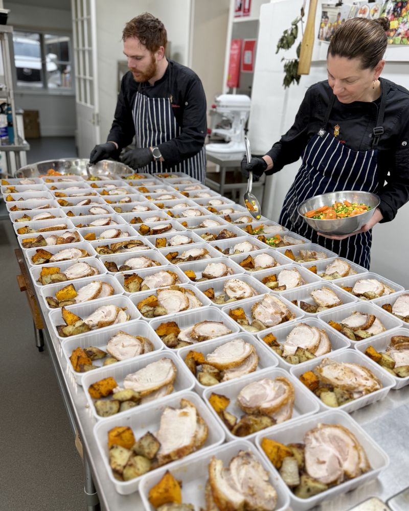 Two people wearing chef uniforms put together roast pork meals in the Government House kitchen