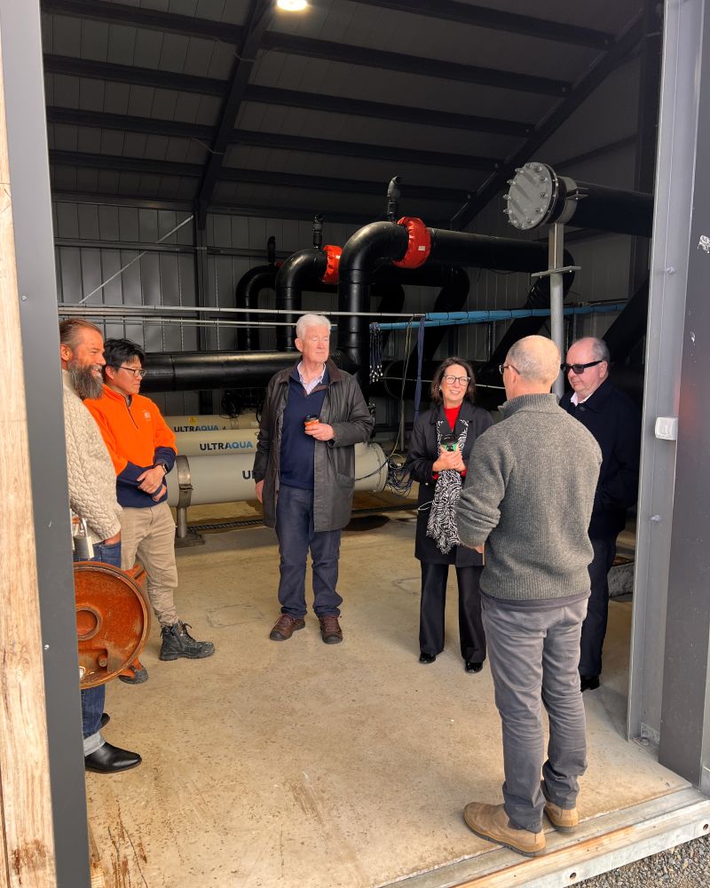 The Governor and Dr Walters stand in a shed with four other men. Machinery is visible in the background
