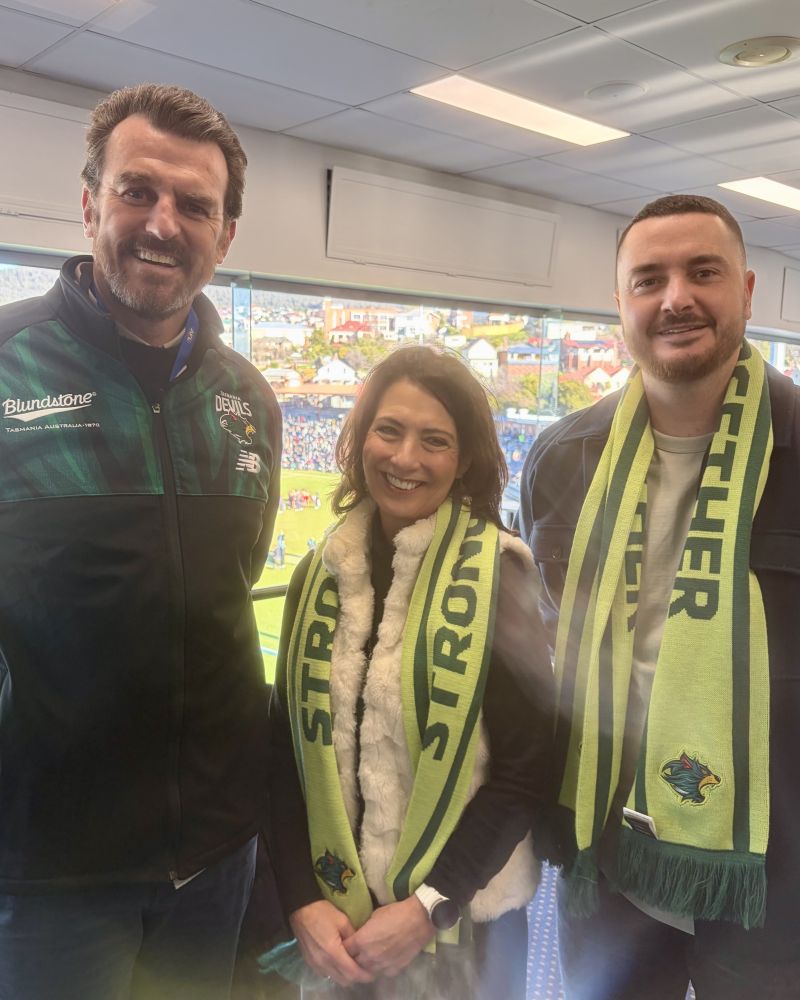 The Governor stands with two men indoor, while a football match takes place outside.