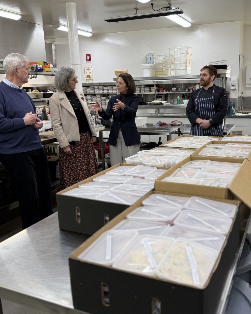 The Governor and Dr Walters speak to a woman in the Government House kitchen. In the foreground prepackaged meals are sitting in boxes.