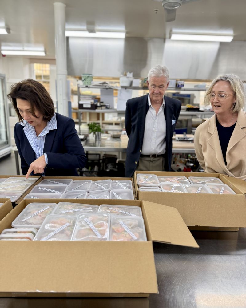 The Governor stands with Dr Walters and a woman in the Government House kitchen, looking at a table full of prepackaged meals