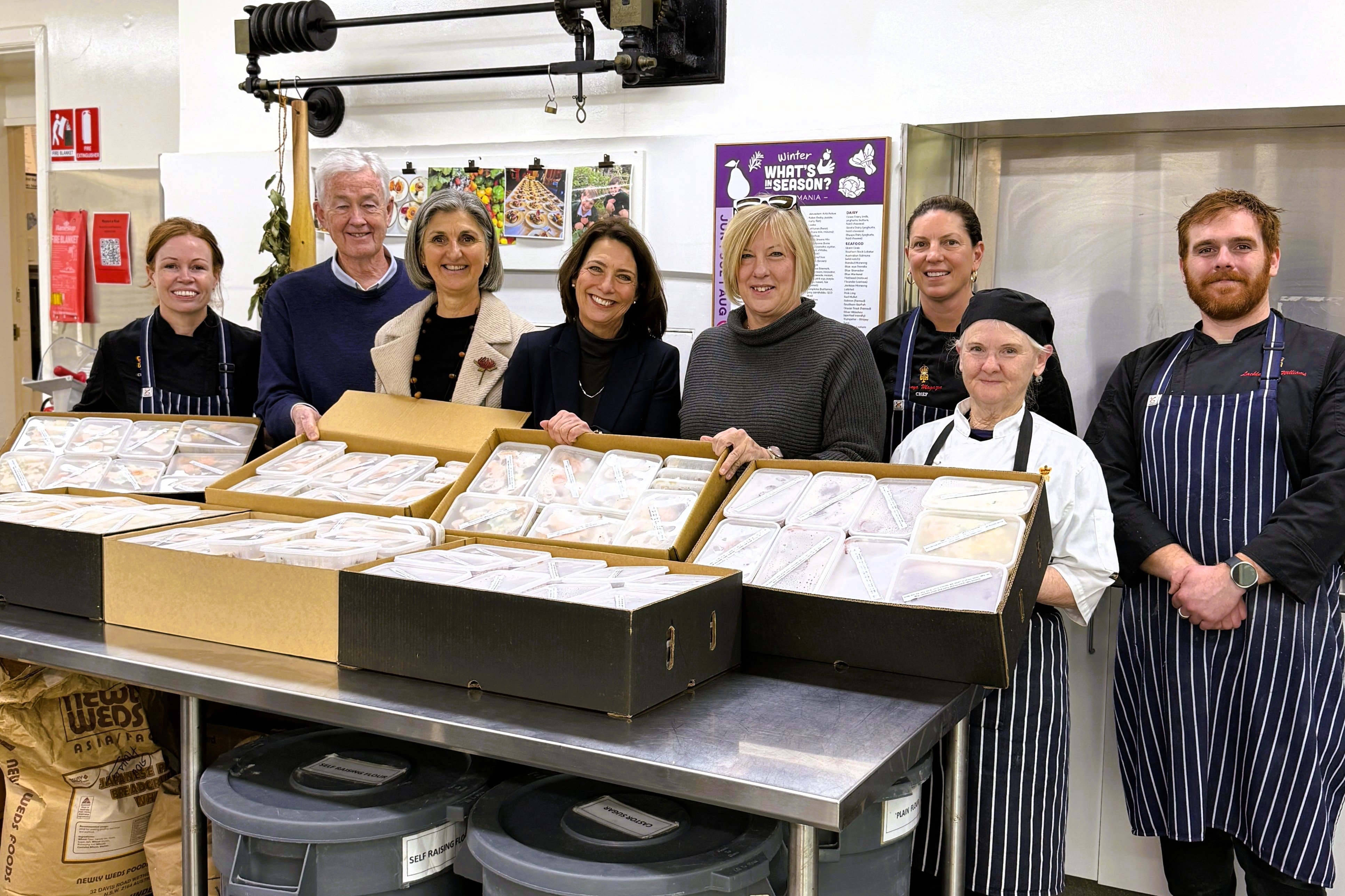 The Governor and Dr Walters stand with six people in the Government House kitchen, holding boxes of prepackaged meals. Everyone is looking at the camera and smiling.
