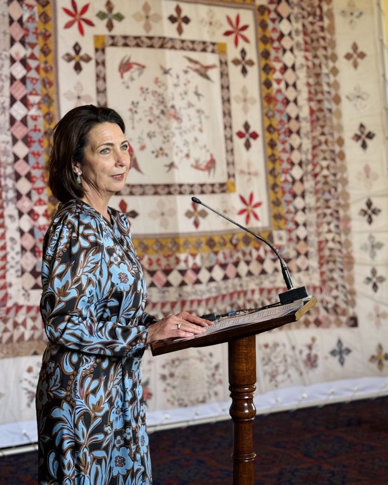 The Governor stands indoors at a lectern, with a colourful quilt on display behind her