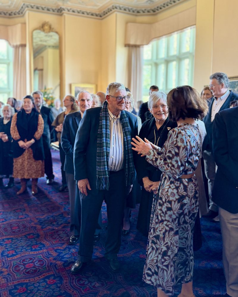 The Governor stands in the Government House Drawing Room speaking to a man and a woman, while several other people mingle with each other behind them